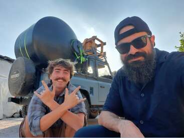 Two men pose cheerfully in front of a rugged vehicle loaded with large black containers. One kneels flashing hand signs, the other wears sunglasses and a beard.
