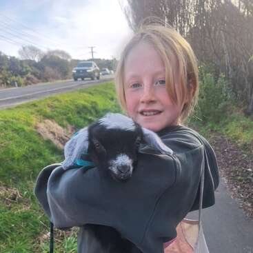 A smiling child with light hair holds a small black and white baby goat while standing on a roadside path, with green grass and trees behind them.