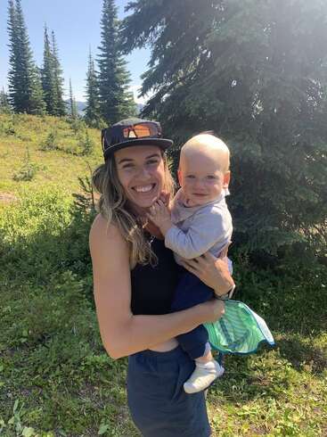 Una mujer sonriente, con gorra y camiseta de tirantes negra, sostiene a un bebé feliz al aire libre. Están entre hierba verde y árboles de hoja perenne en un día soleado.