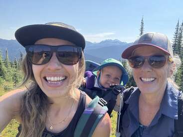 Tres generaciones disfrutan juntas de una soleada excursión por la montaña. Dos mujeres con gafas de sol sonríen mientras un bebé en una mochila portabebés sonríe, rodeado de pintorescos bosques y picos lejanos.