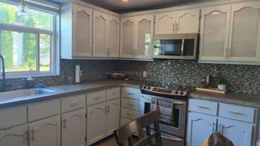 Esta cocina cuenta con armarios blancos, electrodomésticos de acero inoxidable, un mosaico de azulejos, una gran ventana sobre el fregadero, encimeras grises y un ambiente acogedor.