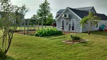 A small, gray house with white trim sits beside a fenced vegetable garden, surrounded by green grass, trees, and outdoor seating under a sunny sky.