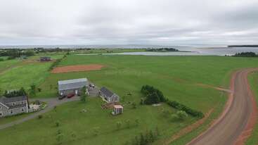 This image shows a rural landscape with a few houses, barns, green fields, a winding dirt road, and a distant body of water under a cloudy sky.