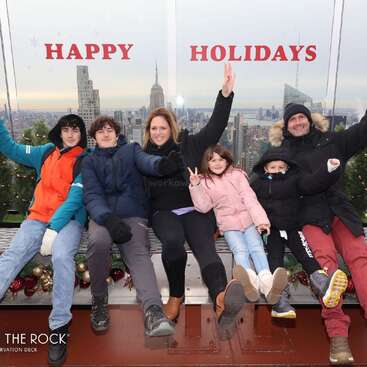 Une famille de six personnes pose joyeusement sur une terrasse panoramique, avec l'inscription « Joyeuses fêtes » en arrière-plan et la silhouette de New York en toile de fond, vêtue de tenues d'hiver.
