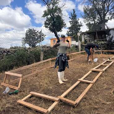 A woman in white boots and gloves poses with thumbs up while working outdoors on a wooden frame structure, with a man and dog in the background.