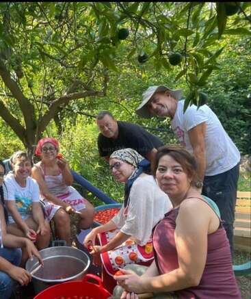 Un grupo de personas está sentado al aire libre bajo un árbol, sonriendo mientras preparan tomates frescos juntos. Parecen felices, compartiendo una actividad en común en un entorno natural.