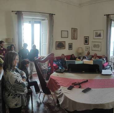 Un grupo de personas se reúne en un salón acogedor y decorado con obras de arte. Algunos están sentados en los sofás, otros de pie, mientras un arpa y un piano acaparan toda la atención.