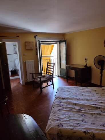 Un acogedor dormitorio con paredes amarillas, suelos de madera, una cama con sábanas de flores, una silla, un escritorio, un ventilador, puertas abiertas que dan a un balcón y un cuarto de baño contiguo.