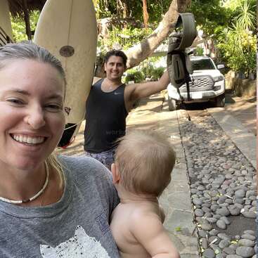 Das Bild zeigt eine Frau mit einem Baby im Arm, hinter ihr ein Mann mit einem Surfbrett und Autoschlüsseln in einer tropischen Umgebung mit Bäumen und einem weißen LKW.
