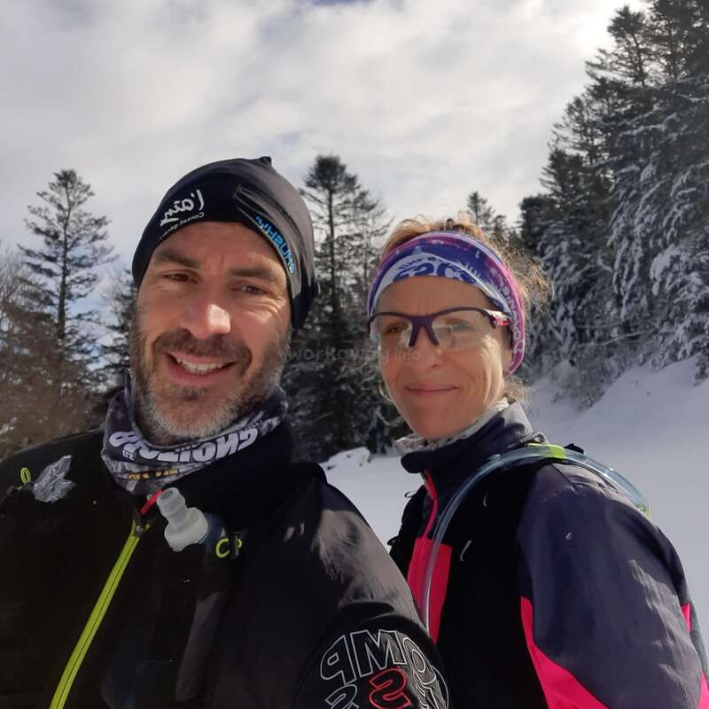 The image shows a man and woman in winter attire, posing for a selfie in a snowy landscape with trees and a cloudy sky in the background.
