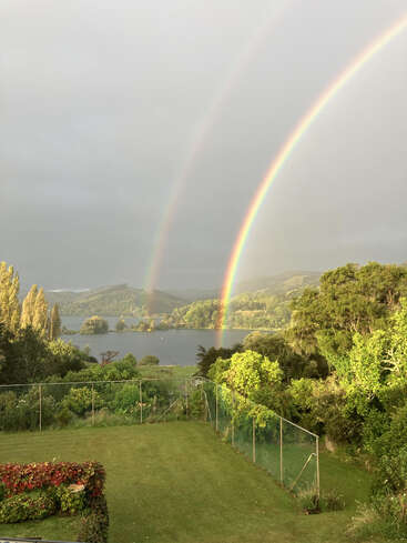 A vibrant double rainbow arcs over a lush, green landscape with manicured lawn, trees, lake, and distant hills under a cloudy sky, creating a serene scene.