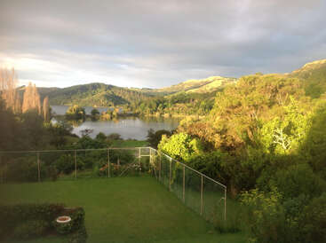 A lush green garden is bordered by a wire fence, overlooking a tranquil lake surrounded by vibrant, sunlit trees and rolling hills under a dramatic sky.