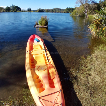 Una kayak de color naranja brillante descansa en la tranquila orilla de un lago, frente a un pequeño muelle. Una exuberante vegetación rodea las tranquilas aguas, bajo un cielo azul claro.