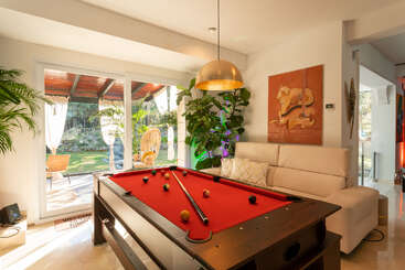 Una moderna sala de estar luminosa cuenta con una mesa de billar roja, un sofá blanco, plantas, obras de arte vibrantes, grandes ventanas y un acogedor patio exterior con sillas de mimbre.