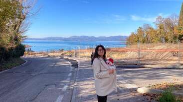 A woman in a beige coat holds a baby wrapped in a blanket. They stand on a quiet road with a scenic lake and mountains in the background.
