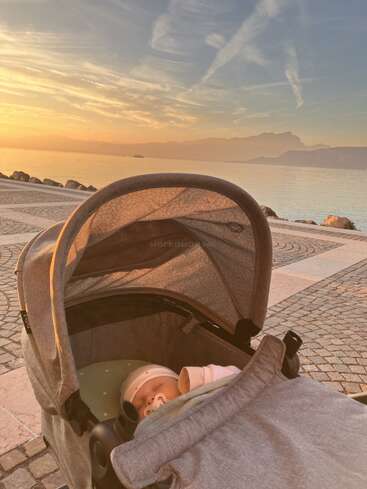 A sleeping baby in a stroller by a peaceful waterfront at sunset, with soft golden light, a calm sea, distant mountains, and beautiful sky. Serene atmosphere.