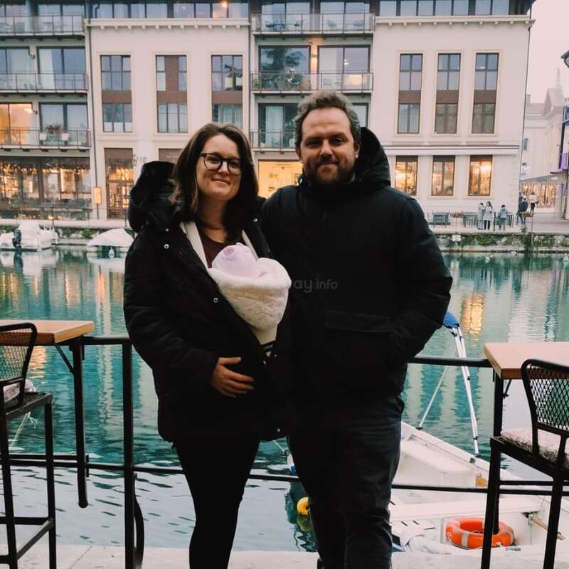 A couple stands together by a waterfront, holding a swaddled baby. Behind them are modern buildings, boats, and reflections on calm blue water. Smiling warmly.