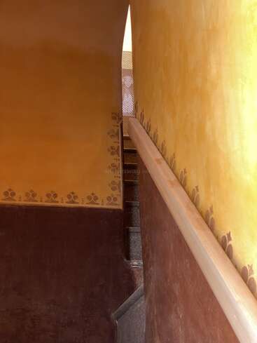A narrow staircase with brown and yellow painted walls, floral border designs, and a wooden handrail. Light shines through a patterned screen at the top.