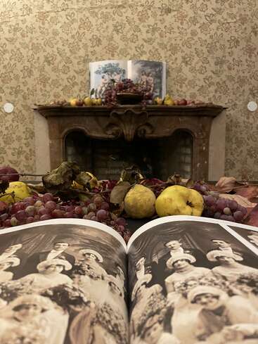 An open book displays a vintage wedding photograph in the foreground. Fruit and grapes decorate a table and mantel, with a fireplace and floral wallpaper behind.