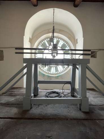 The image shows an old clock mechanism inside a tower. The clock face is visible through the arched window behind wooden and metal machinery parts. Cables rest below.