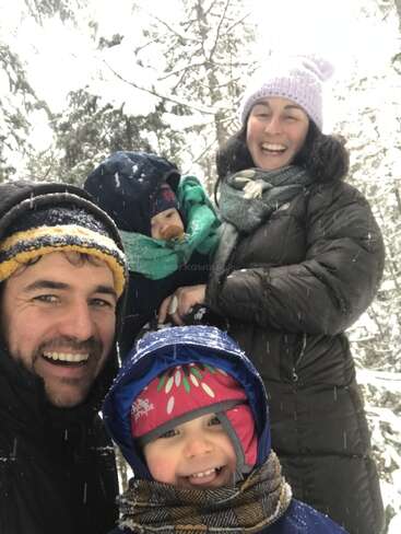 A family of four, bundled in winter clothes, smiles joyfully outdoors amid falling snow and snowy trees, capturing a happy, playful moment in a winter wonderland.