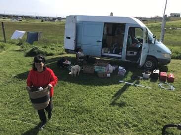 A woman in a red sweater carries a basket across a grassy field. Behind her, a van is open, belongings and a dog are scattered nearby.