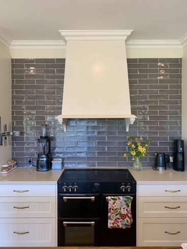 This kitchen features a black stove, white cabinetry, gray subway tile backsplash, blender, coffee maker, kettle, vase with yellow flowers, hanging utensils, and a floral towel.