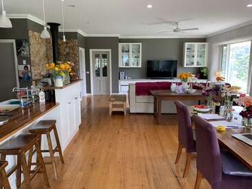 This open-plan living space features a wooden floor, kitchen, cozy lounge, dining area, vibrant flowers, natural light, and modern decor, creating a warm, inviting atmosphere.