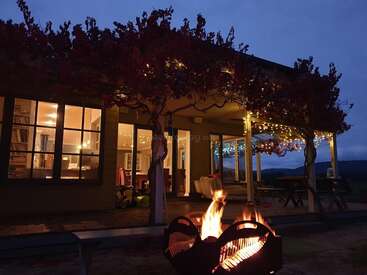 A cozy house at dusk, warmly lit inside, with string lights on the porch, surrounded by trees and a glowing fire pit in the foreground, inviting relaxation.