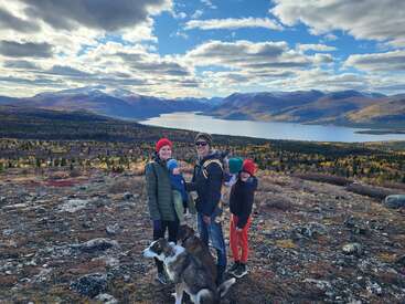 A family with two children and a baby, along with a dog, stands on a rocky hilltop, overlooking a vast lake and mountainous landscape.