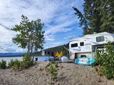 A scenic lakeside campsite with an RV, paddleboard, picnic table, and laundry drying. Surrounded by trees, mountains, and blue sky with picturesque clouds. Relaxing atmosphere.