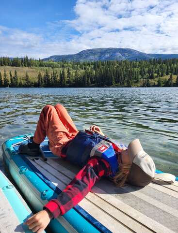 A child relaxes on a paddleboard, wearing a life jacket and cap, floating on a tranquil lake with forested hills and mountains under a bright blue sky.