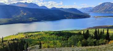 A serene landscape features a large blue lake, lush green forests, distant mountains under a bright blue sky with scattered clouds, and vibrant sunlight illuminating the scene.