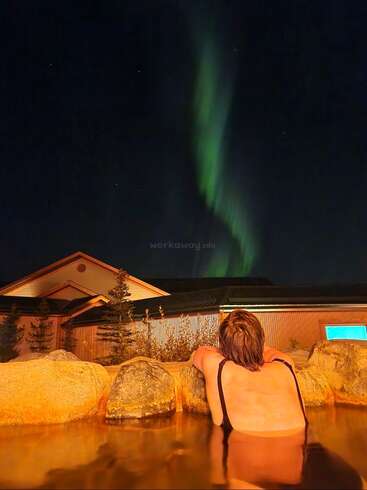 A person relaxes in a hot spring, gazing at the spectacular green Northern Lights illuminating the night sky above a cozy cabin surrounded by rocks.