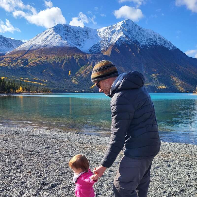 A man and small child walk along a rocky lakeshore, with turquoise water, forest, and majestic snow-covered mountains under a clear blue sky. Serene nature moment.