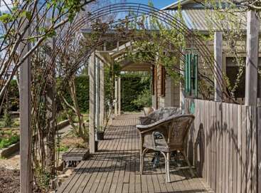 Une terrasse rustique en bois, où trône une chaise en osier, se trouve sous une arche feuillue, à côté d'une charmante maison. La végétation luxuriante et la lumière naturelle créent une ambiance paisible.