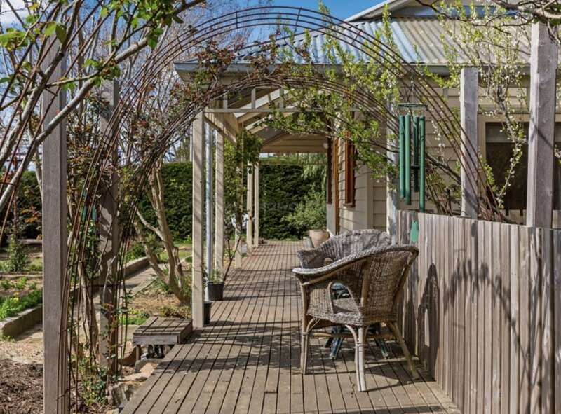 Une terrasse rustique en bois, où trône une chaise en osier, se trouve sous une arche feuillue, à côté d'une charmante maison. La végétation luxuriante et la lumière naturelle créent une ambiance paisible.