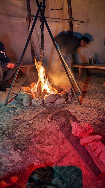 Zwei Menschen sitzen um ein warmes, helles Lagerfeuer in einem Zelt. Ein Dreibein stützt eine hängende Kette über den Flammen. Gemütliche, rustikale und einladende Atmosphäre.