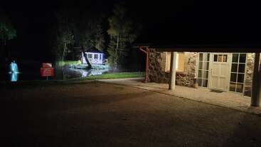 Ein Steingebäude mit Säulen ist nachts beleuchtet. Im Hintergrund spiegeln sich ein beleuchteter Pavillon und eine Wasserfontäne auf einem ruhigen Teich, der von Bäumen umgeben ist.