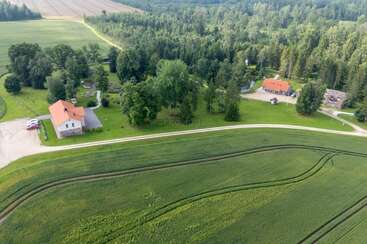 Dieses Luftbild zeigt eine ländliche Landschaft mit grünen Feldern, einer gewundenen Schotterstraße, Häusern mit roten Dächern, Autos und dichtem Wald im Hintergrund.