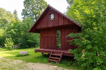 A small red wooden cabin with a porch and stairs sits surrounded by lush green trees and grass, evoking a peaceful, rustic forest retreat.