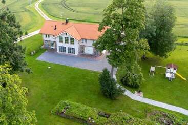 Aerial view of a countryside house with a red roof, surrounded by expansive green lawn, large trees, a playground, a driveway, and open farmland.