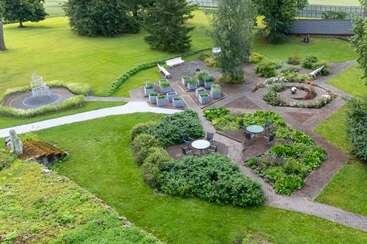 A beautifully landscaped garden with pathways, seating areas, tables, benches, lush greenery, flowerbeds, a small water fountain, and decorative plants in a serene outdoor setting.