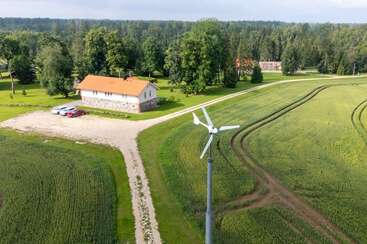 A countryside scene features a wind turbine, a house with an orange roof, parked cars, lush green fields, winding paths, trees, and a distant forest background.