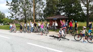A group of cyclists, including adults and children, pose cheerfully beside a road. They raise their arms, with trees, greenery, and a small shelter in the background.