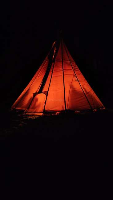A glowing orange teepee stands illuminated in complete darkness. The light from within casts shadows on the tent, creating a cozy, inviting, and mysterious atmosphere.