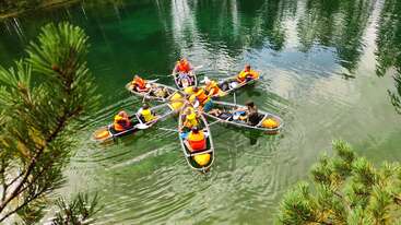 A group of people in life jackets sit in six canoes arranged in a star shape on a clear green lake, surrounded by trees and nature.