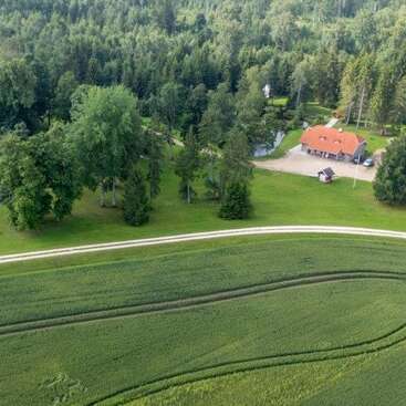 Esta imagen aérea muestra un paisaje rural con dos casas, una carretera en curva, campos de cultivo, varios árboles, coches aparcados y un denso bosque de fondo.