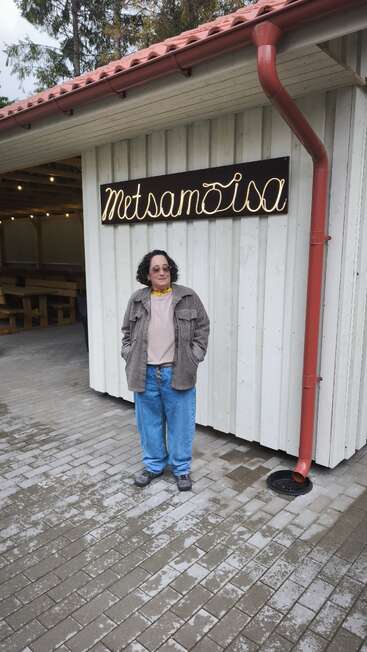 Eine Person mit lockigem Haar, die eine Sonnenbrille und Freizeitkleidung trägt, steht vor einem weißen Holzgebäude mit einem Schild „Metsamoisa“ und einem roten Fallrohr.
