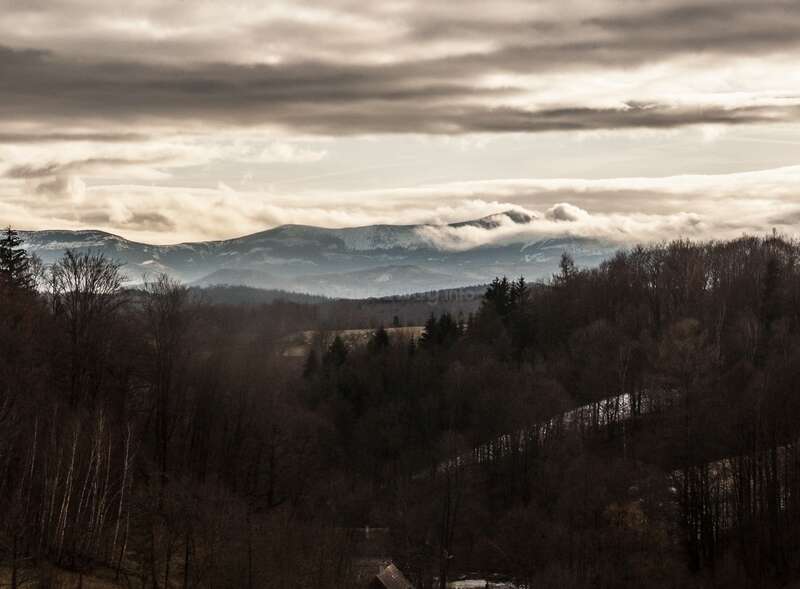 The image depicts a serene landscape featuring a mountain range in the distance, with trees and hills in the foreground, set against a cloudy sky.